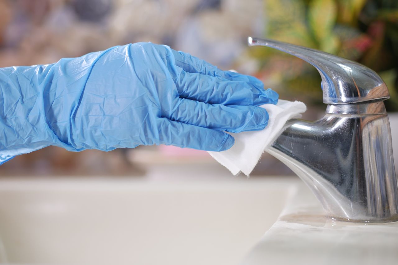 Portfolio Close-up of gloved hand wiping a stainless steel faucet with a cloth indoors.