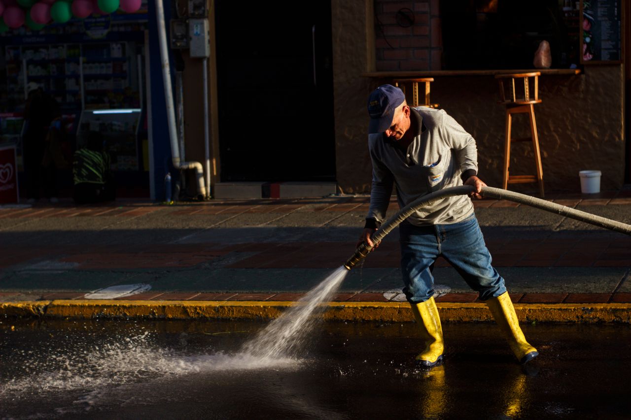 Portfolio A man using a water hose to clean a city street, wearing yellow rain boots and a cap.