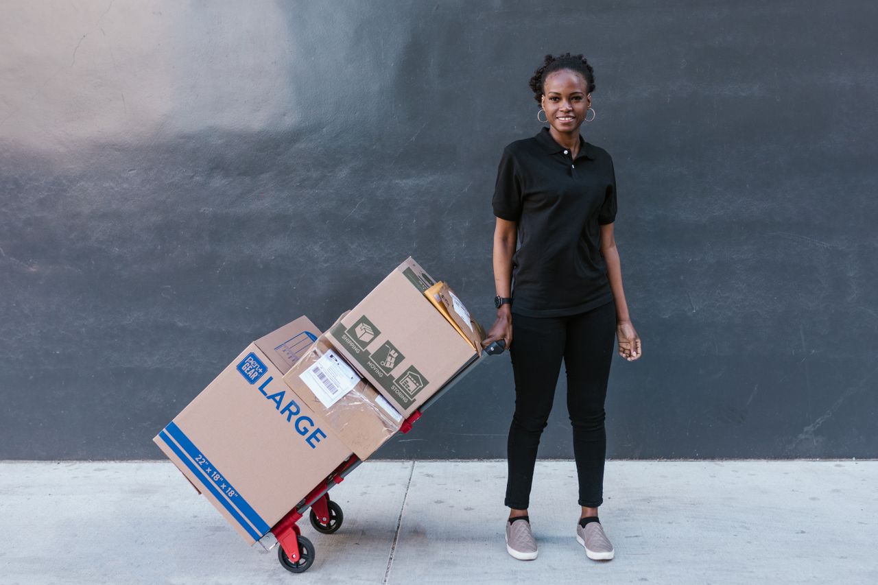 About Black woman in uniform pulling a dolly with large boxes, photographed outdoors against a dark wall.